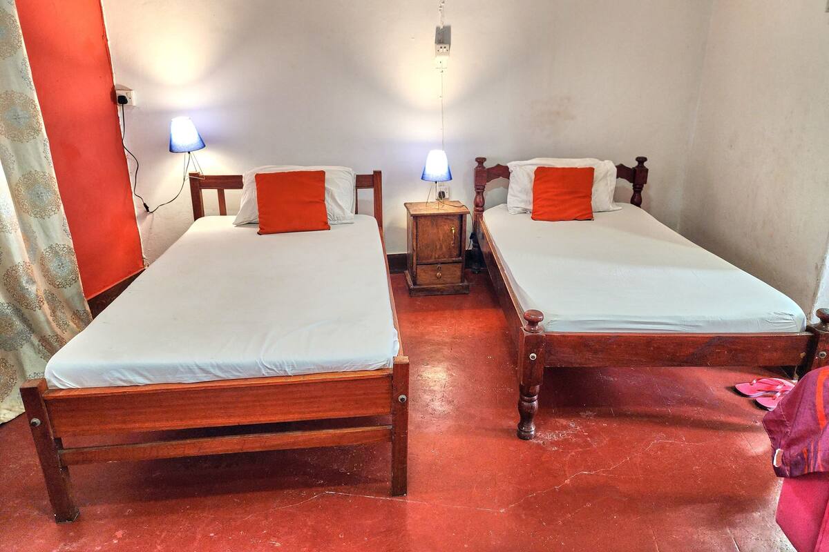 Una habitación con dos camas de madera, cada una con ropa de cama blanca y almohadas naranjas. Hay lámparas de noche sobre mesitas de madera.