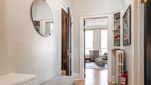 A hallway with a bench, a round mirror, and a door leading to a living area with a radiator, ottomans, and a fire extinguisher.