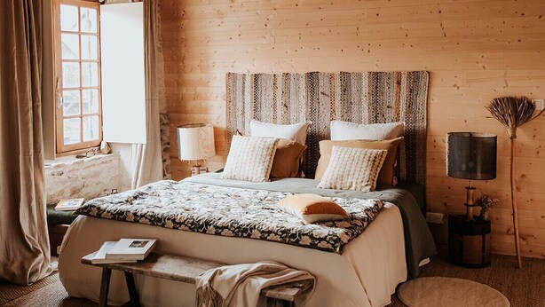 Een rustieke slaapkamer met houten vloer, een groot bed, een bankje, een nachtkastje en een raam met gordijnen.