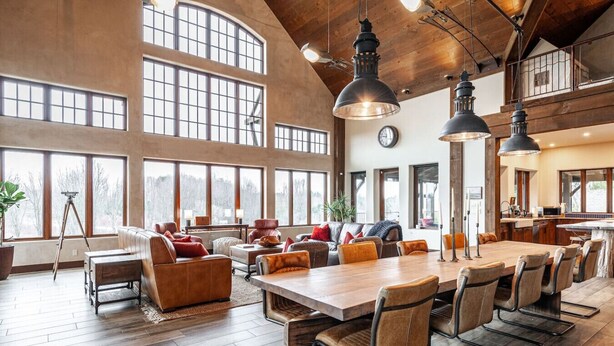 A spacious dining area with a long wooden table and multiple chairs, large windows, and a high ceiling with exposed beams. - The Elmwood Wellness Estate (Dundas)