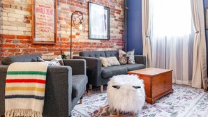 Un salon chaleureux avec un mur en briques, deux canapés gris, un tabouret moelleux blanc et une table basse en bois.