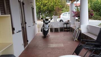 Een overdekt terras met daarbuiten een scooter, witte stoelen en een tafel.