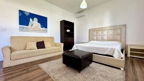 A room with a beige sofa, a black ottoman, a bed with a white headboard, and a painting of a Buddha on the wall. - Villetta Certosina 2 (Racale)