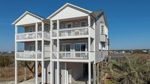 A two-story beachfront house with a balcony on each floor, a covered entryway, and a sandy beach in the background. - Ocean Heir -The Water is Calling (North Topsail Beach)