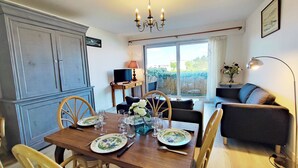 Un comedor con una mesa de madera dispuesta para cuatro personas, rodeada de sillas. Un televisor está sobre un mueble a la izquierda, y un sofá al fondo.