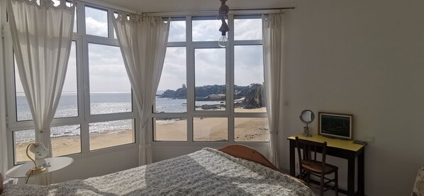 A bedroom with a large window overlooking a beach, a bed, a desk with a chair, and a small table with a lamp.