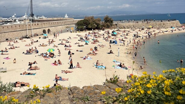 A beach with people sunbathing and swimming, a stone wall, and boats in the background.