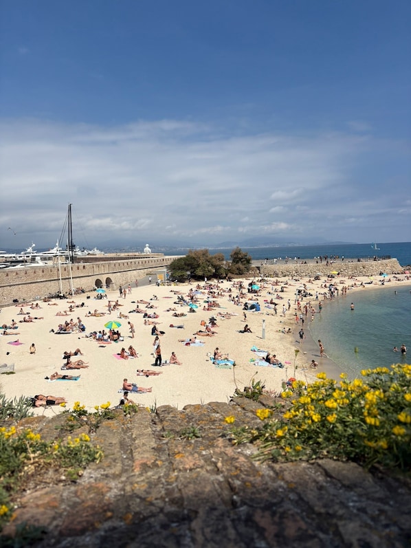 A beach with people sunbathing and swimming, a stone wall, and boats in the background.