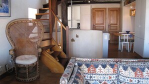 A cozy room with a wicker chair, a patterned sofa, a wooden staircase, and a kitchen area with cabinets and a refrigerator.