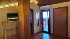 Une pièce aux murs recouverts de lambris en bois, un téléviseur posé sur un meuble, une porte donnant sur un espace extérieur et un ventilateur de plafond.