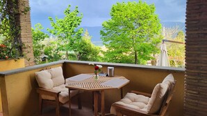 Un balcon avec une table en bois et deux chaises rembourrées, donnant sur un jardin luxuriant et des montagnes lointaines.
