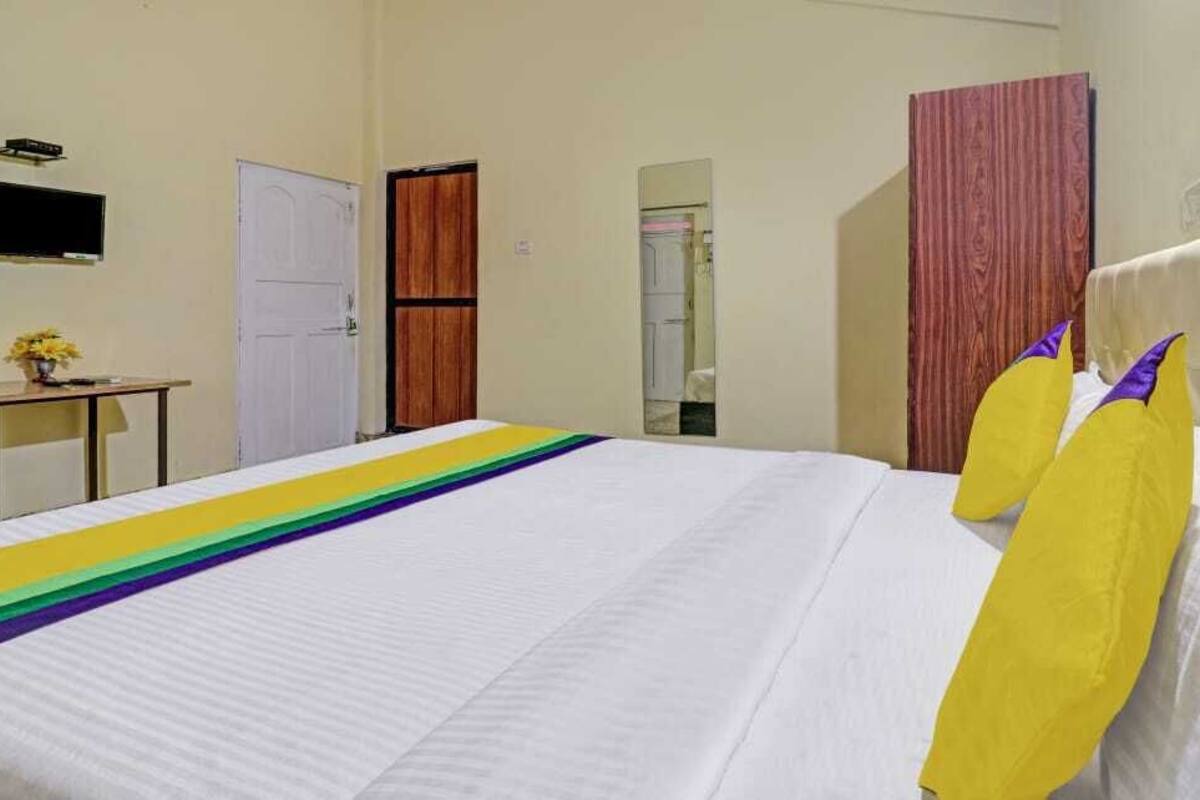 A hotel room with a bed, a wooden wardrobe, a small table with a vase, and a mirror on the wall.