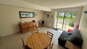 A living room with a wooden dining table and chairs, a sofa, a TV, and a sliding glass door leading to a garden.