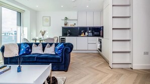 A modern living room with a blue sofa, a white coffee table, and a view of the city through large windows. - Cosy London Apartment Near River Thames (London)