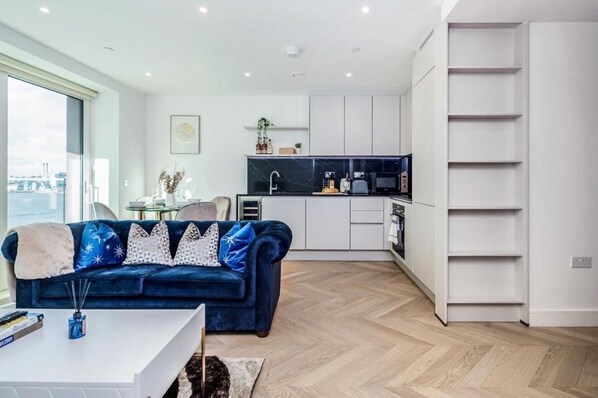A modern living room with a blue sofa, a white coffee table, and a view of the city through large windows. - Cosy London Apartment Near River Thames (London)