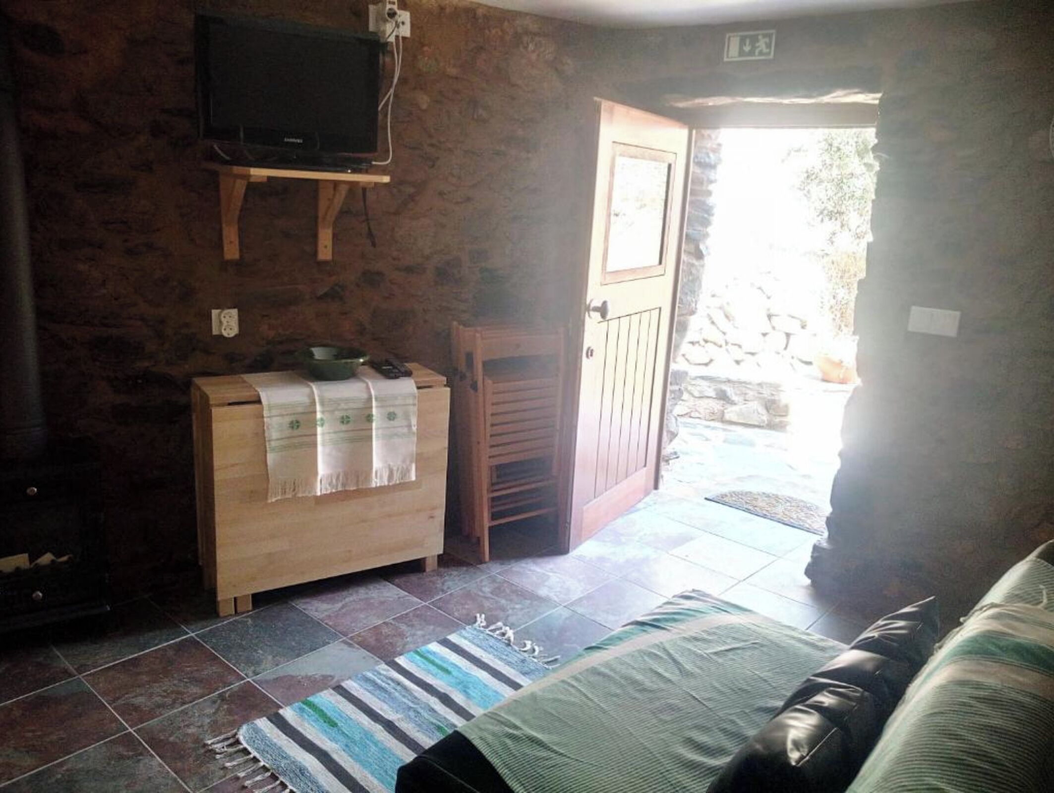 A cozy room with a television on a wooden stand, a small wooden cabinet with a cloth, a door leading outside, and a striped rug on the floor.