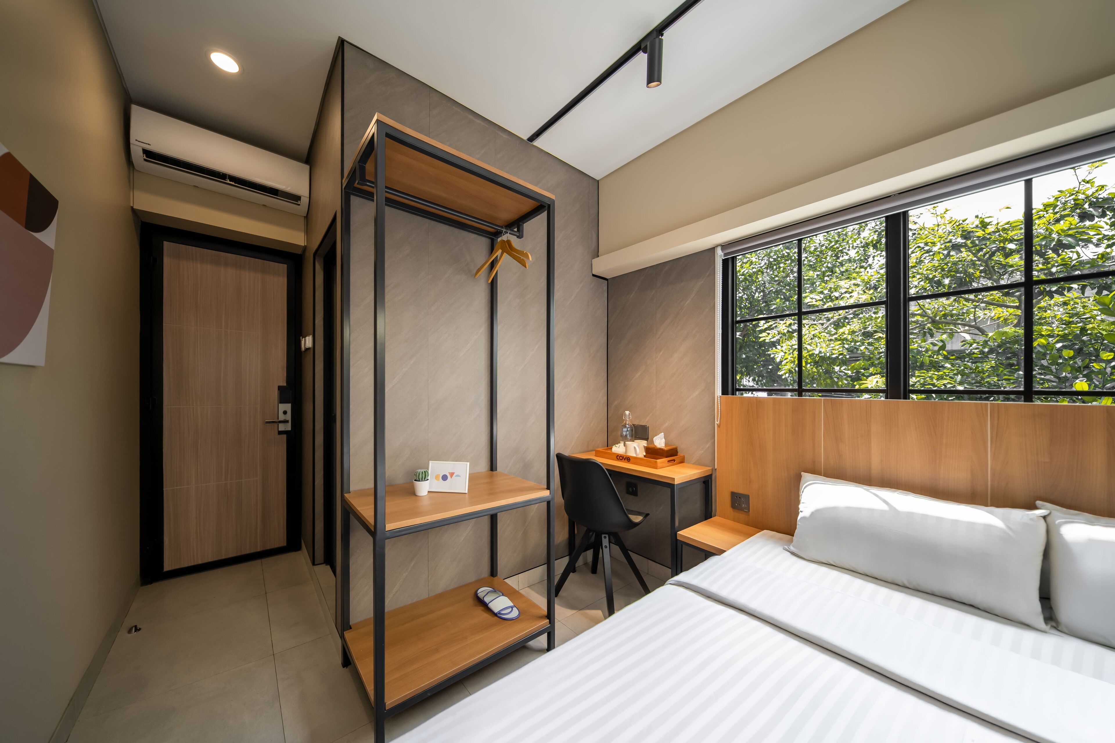 Une chambre d’hôtel moderne, équipée d’un lit, d’un bureau et d’une chaise. Une fenêtre offre une vue sur les arbres.
