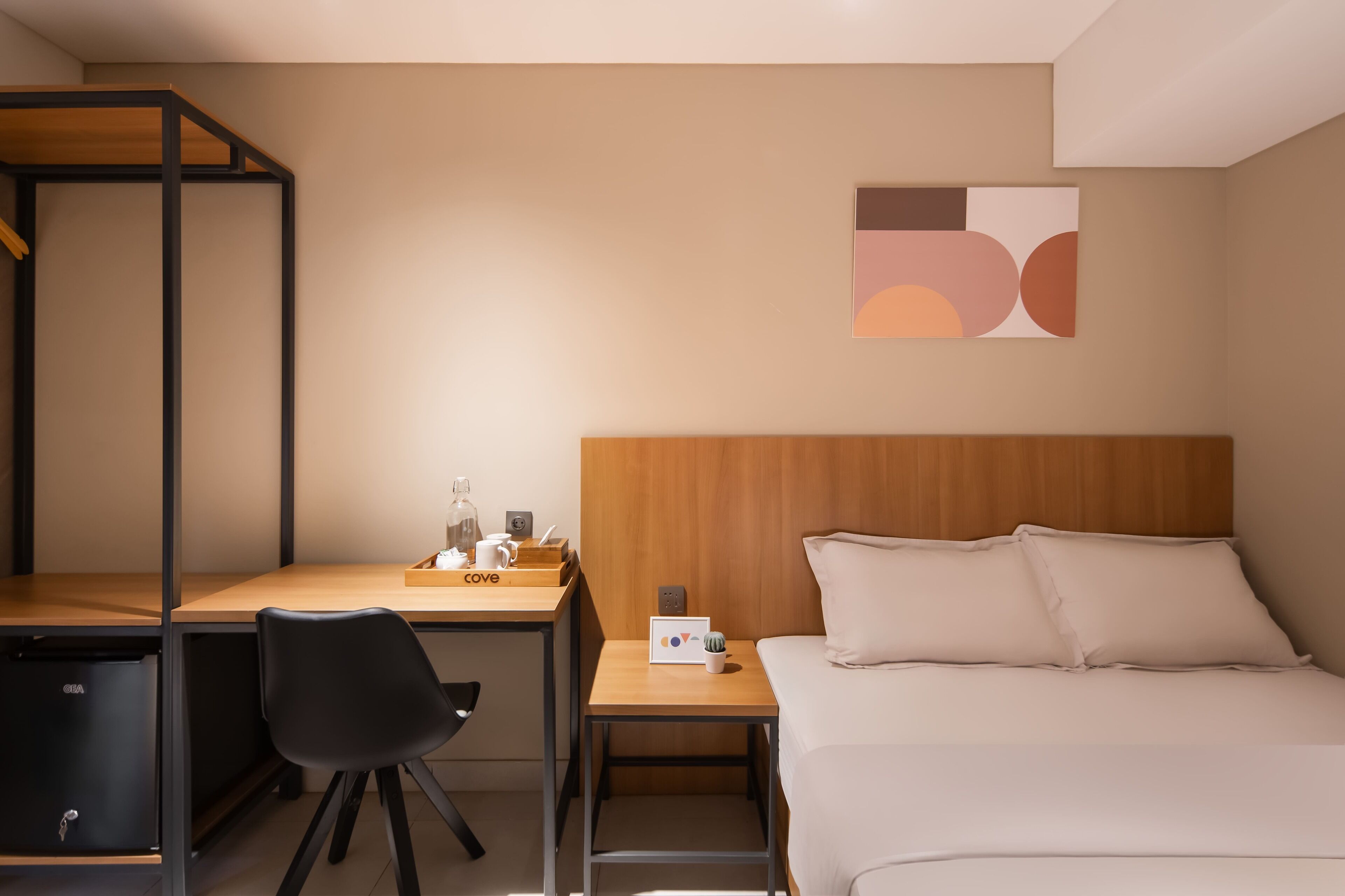 A hotel room with a bed, a desk, a chair, a small table with a lamp, and a wall art piece.