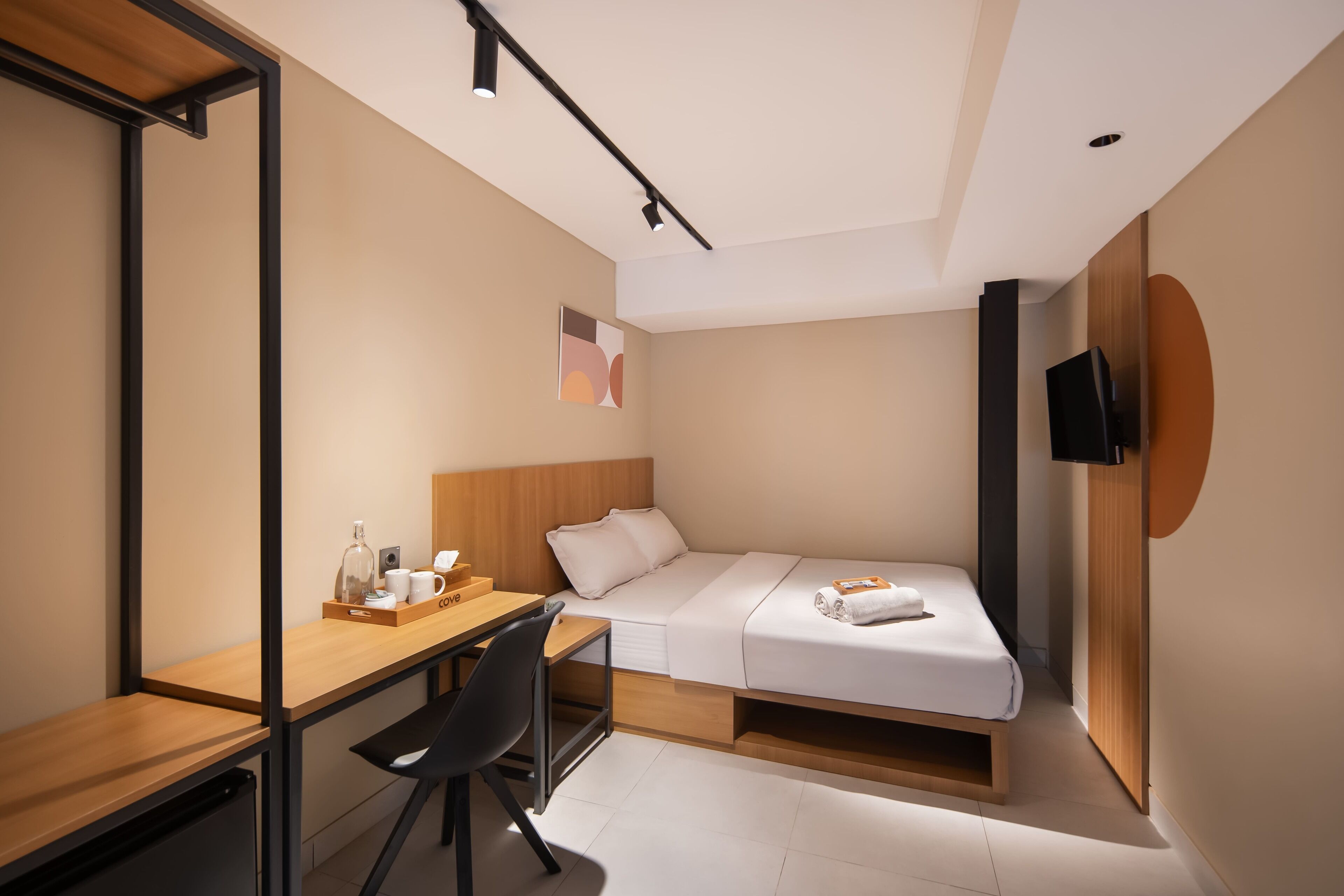 A compact hotel room with a bed, a desk, a chair, a TV, and a small table with a bottle and a cup.