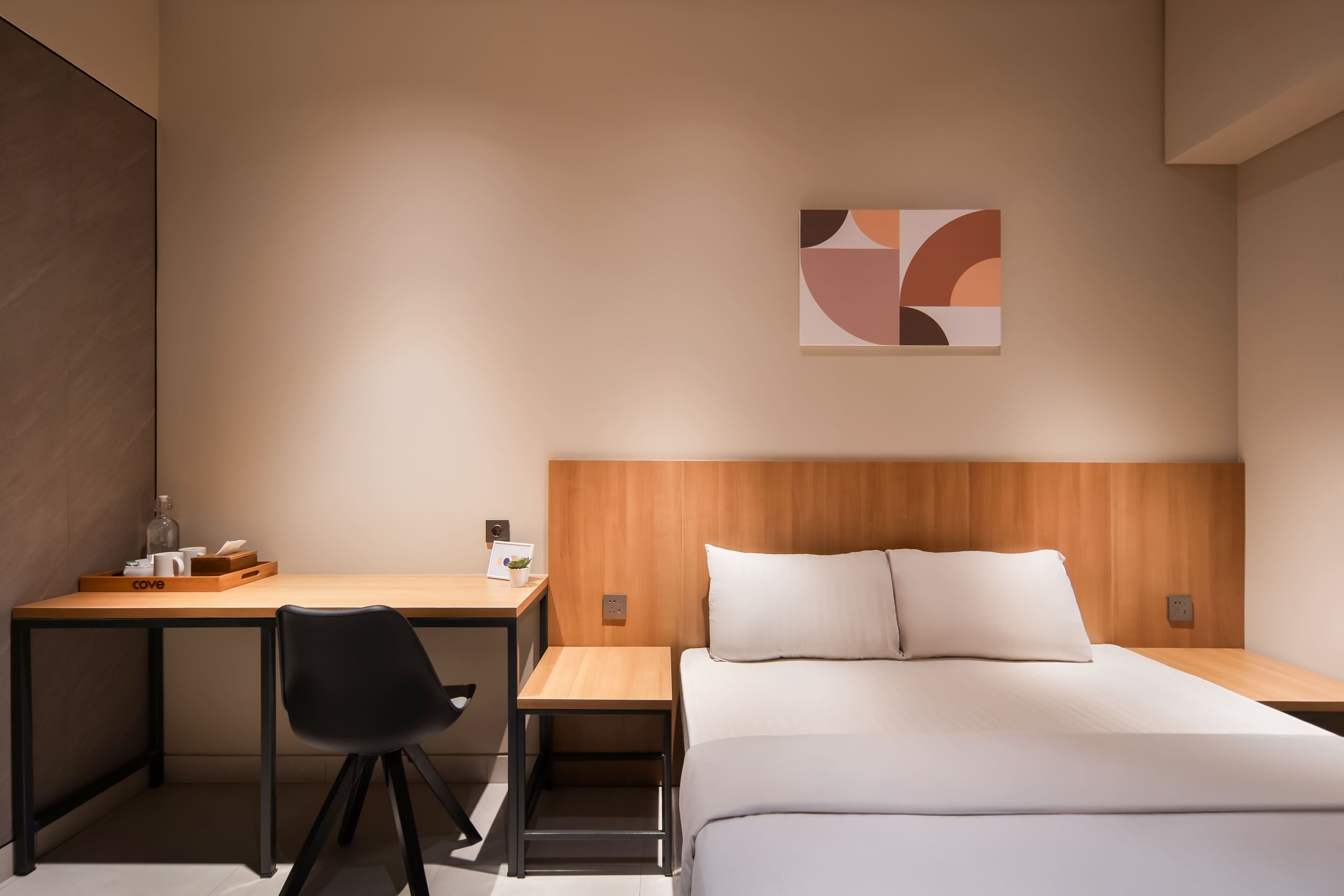 Habitación de hotel con cabecera de madera, cama con ropa blanca, escritorio con silla y una mesita con una botella y una taza.