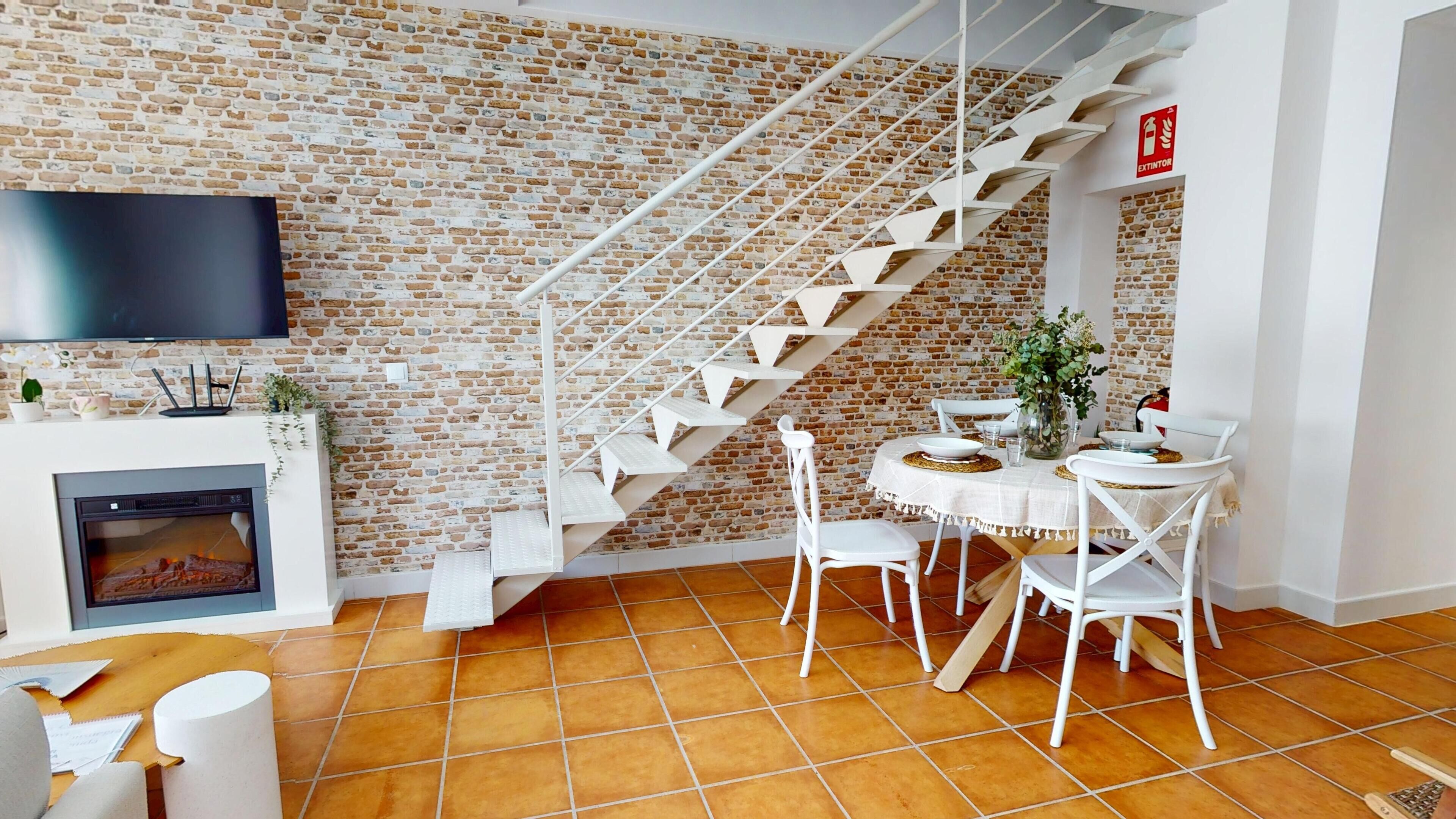 Una sala de estar moderna con pared de ladrillo, chimenea, televisor y zona de comedor con mesa y sillas.