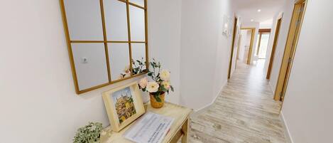 A hallway with a wooden floor, a mirror, a framed picture, a vase with flowers, and a small table with a sign.