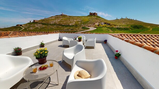 A rooftop terrace with white chairs, a round table with drinks and fruit, and potted plants.
