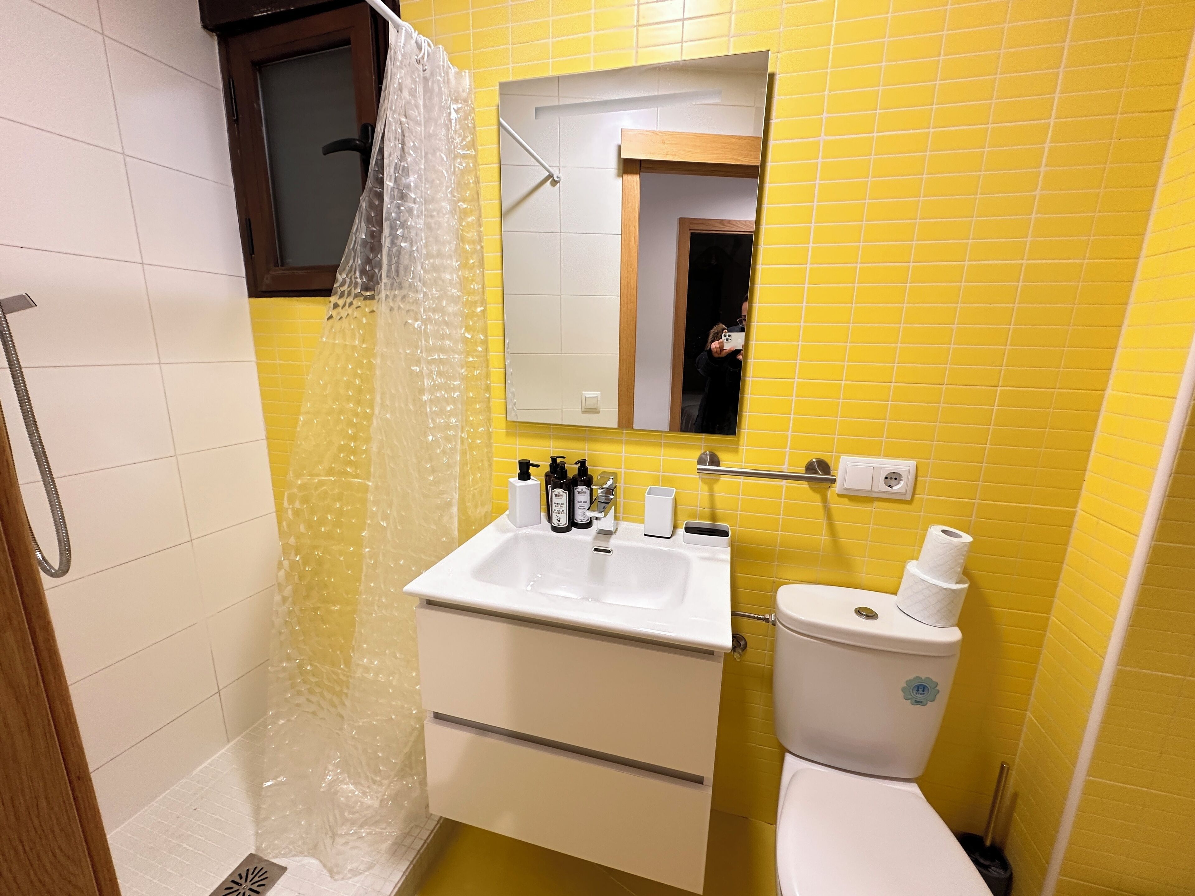 Un baño con pared de azulejos amarillos, un lavamanos blanco con dispensadores de jabón, un inodoro y una ducha con cortina transparente.