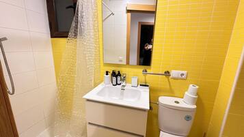 Un baño con pared de azulejos amarillos, un lavamanos blanco con dispensadores de jabón, un inodoro y una ducha con cortina transparente.