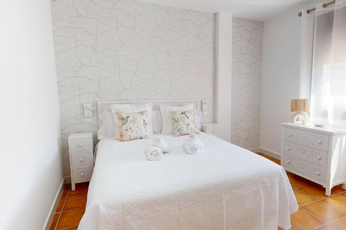 A neatly arranged bedroom with a large bed, a bedside table, a dresser, and a window with curtains.
