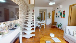 A modern living room with a white sofa, a dining table with chairs, a staircase with a brick wall, and a tiled floor.