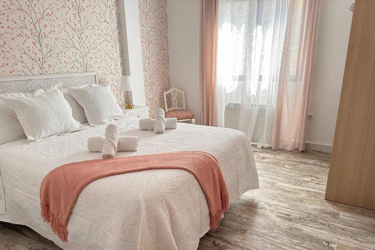A neatly made bed with white linens and pink towels, a wooden floor, and floral wallpaper.