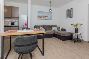 A modern living room with a sectional sofa, a dining table with a chair, a small side table with a vase, and a kitchen area with a microwave and refrigerator. - Apartment Mutak (Vir)