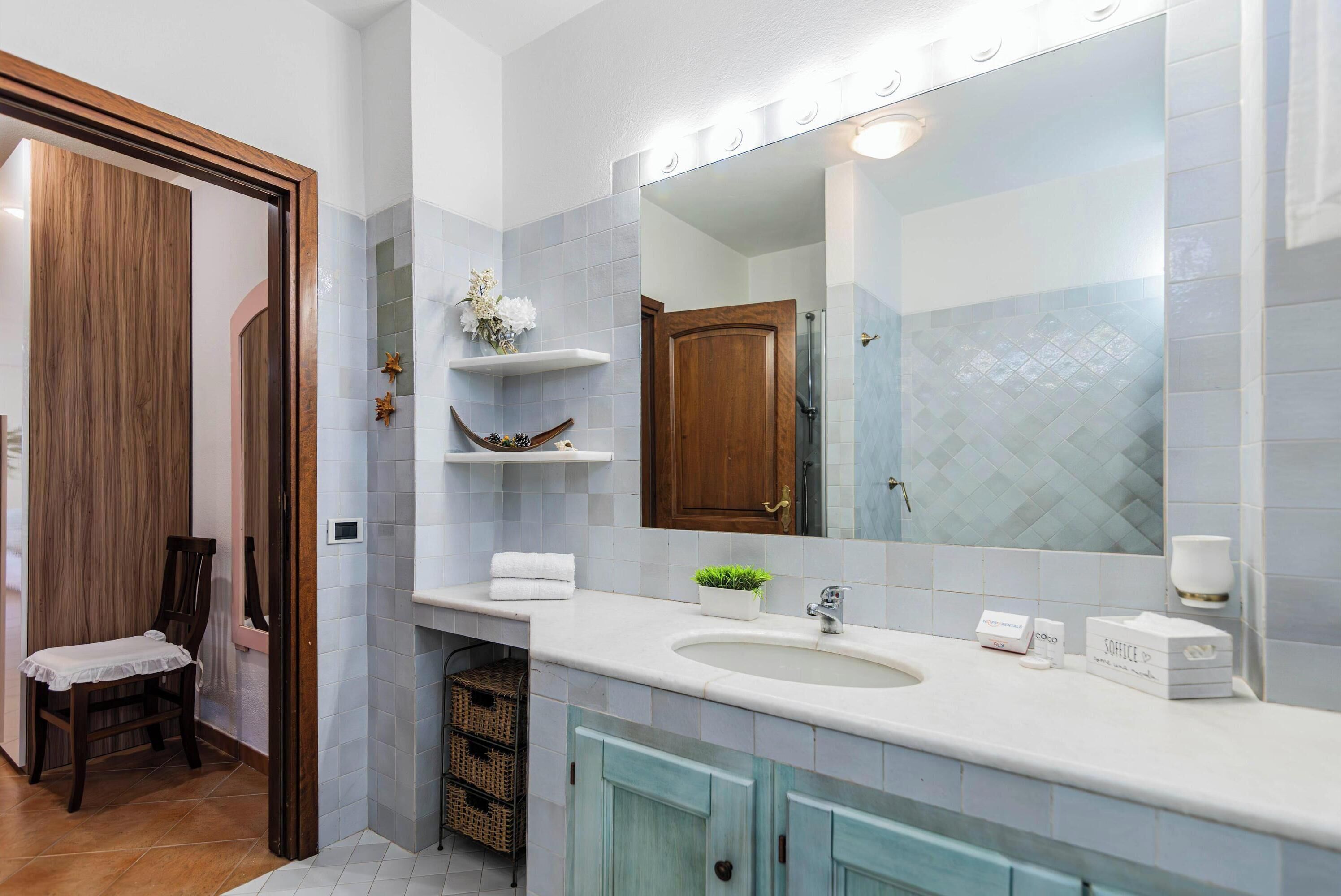 A bathroom with a large mirror, white sink, and blue cabinets. There are shelves with toiletries and a chair.