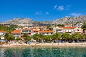 Un pueblo costero con edificios a lo largo del paseo marítimo, un cielo azul despejado y montañas al fondo.