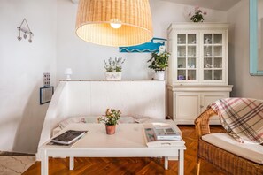 Ett mysigt vardagsrum med en vit soffa, en fåtölj i rotting, ett vitt bord med böcker och en krukväxt, en lampa i rotting och ett vitt skåp med glasdörrar.