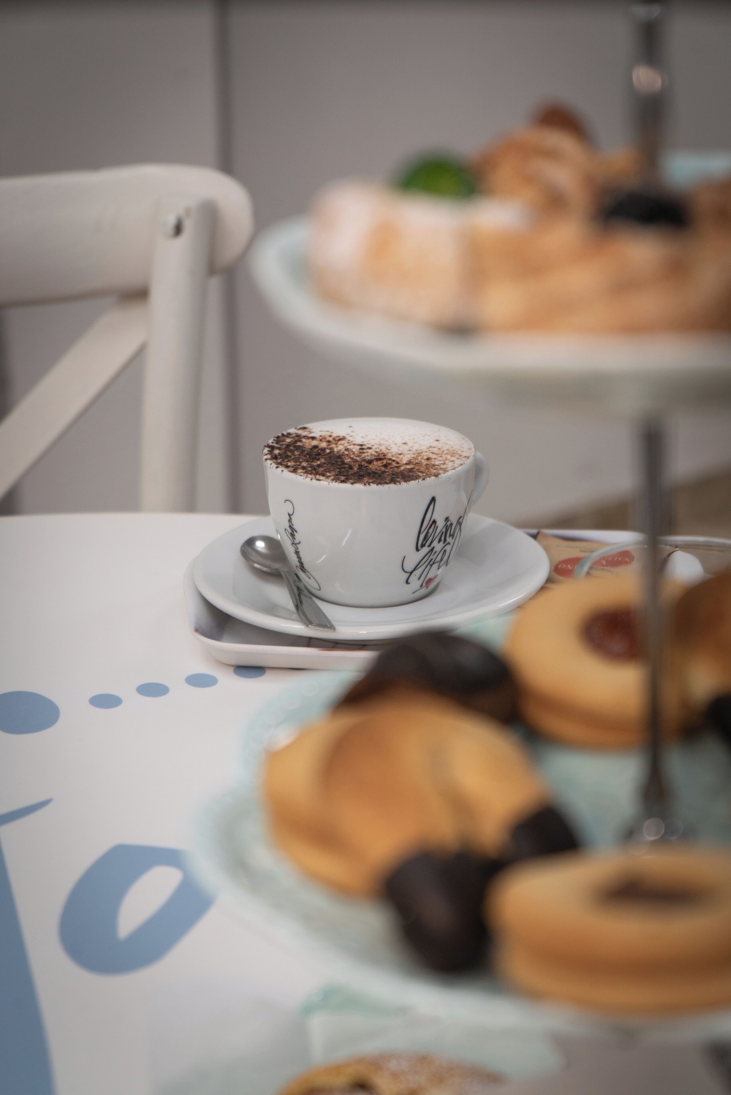 A white cup with a spoon on a saucer, a plate with pastries, and a blurred background with a cake on a stand.