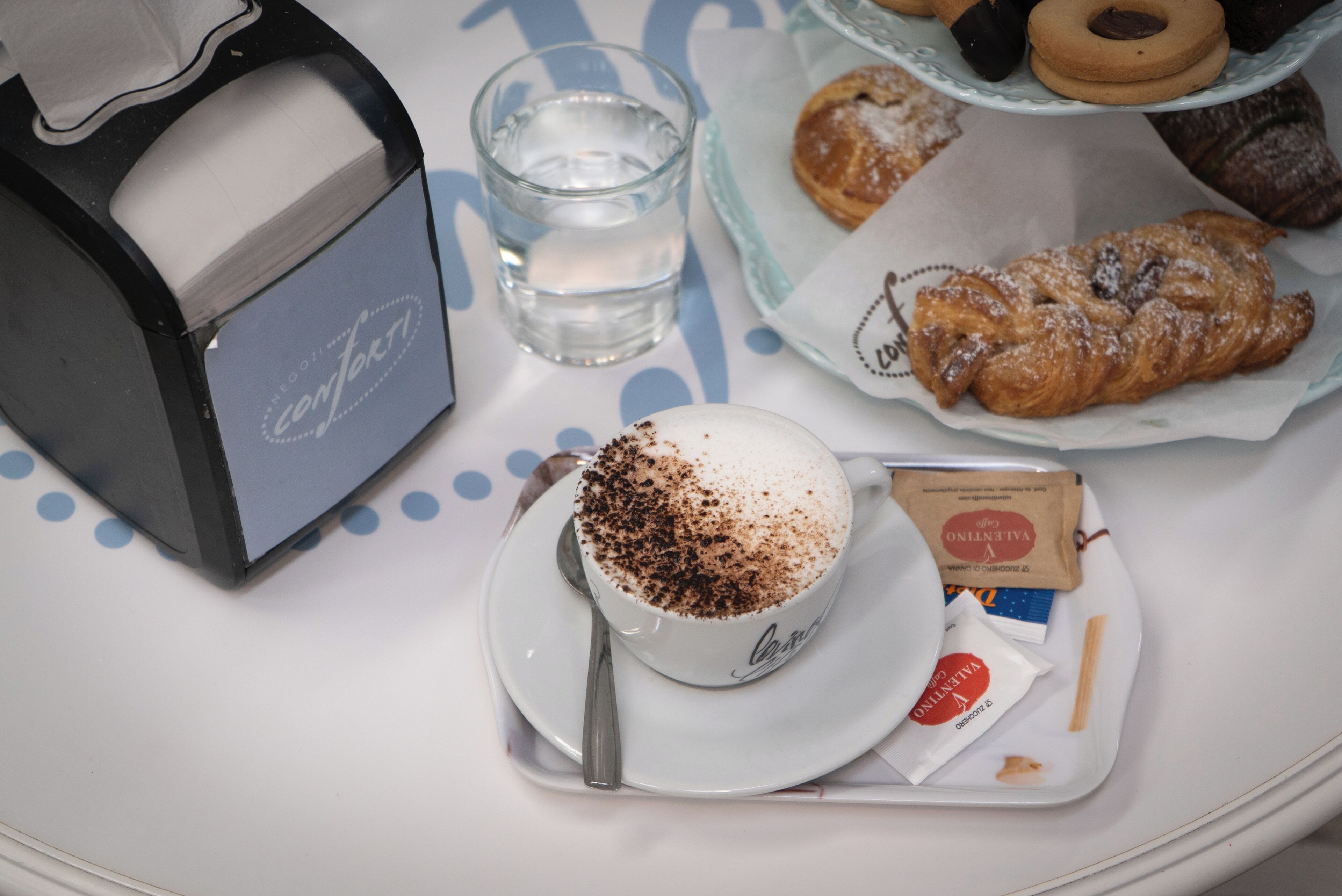 A tray with a cup of coffee topped with cocoa powder, a glass of water, and pastries on a table.