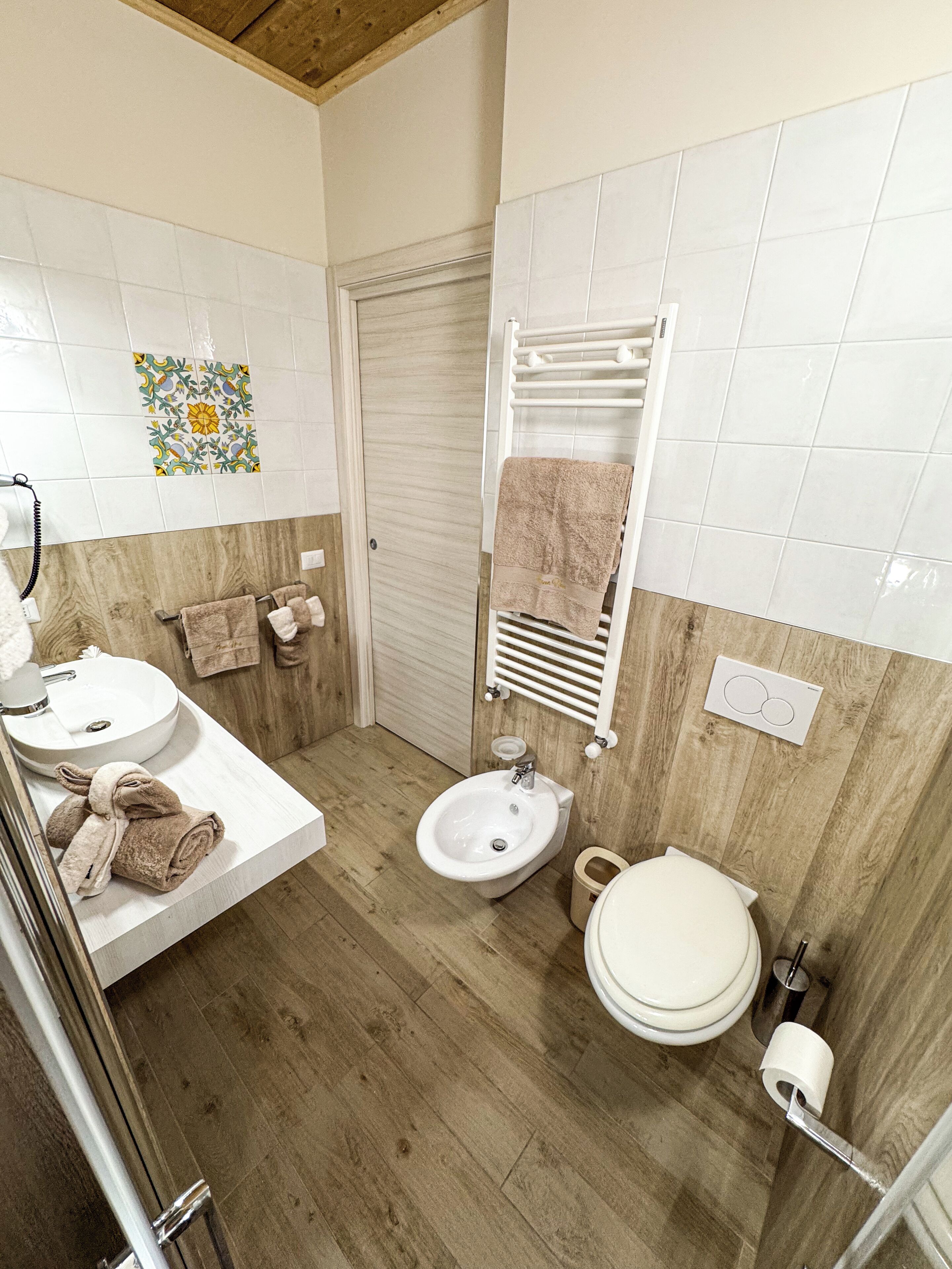 A bathroom with a wooden floor, white tiles, a sink, a bidet, a toilet, and a towel rack.