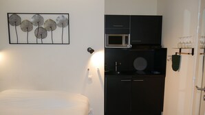 A compact room with a bed, a kitchenette, and a wall-mounted light fixture. - Just 10 minutes from the town center, this cocooning studio is sure to make your stay a pleasant one! (Mulhouse)