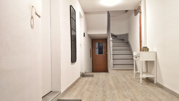 A hallway with a wooden floor, a white door, a staircase, and a small white shelf with a vase. - Just 10 minutes from the town center, this cocooning studio is sure to make your stay a pleasant one! (Mulhouse)