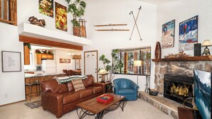 A cozy living room with a fireplace, a sofa, a coffee table, and a television. There is a kitchen in the background with a dining area. - Lemmon Lodge #30 – Lake Access with private sandy beach (Grand Lake)