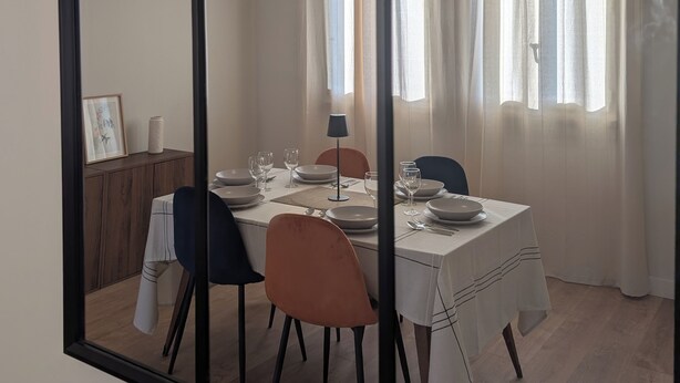 Un coin repas avec une table dressée pour quatre personnes, des chaises, un luminaire suspendu et une fenêtre avec des rideaux.