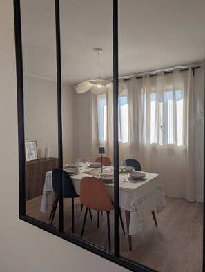 Un comedor con una mesa dispuesta para cuatro personas, sillas, una lámpara colgante y una ventana con cortinas.