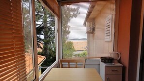 A small kitchen with a table and chair, a window with blinds, and a view of a beach and trees. - Seafront Apartments Pretner (Orebic)
