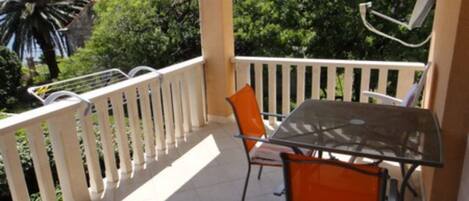 A balcony with a glass table and two orange chairs, surrounded by a white railing and trees.