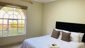 A neatly made bed with a white duvet and two patterned pillows, a window with a view of greenery, and a black headboard.