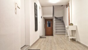 A hallway with a wooden floor, a white door, a staircase, and a small white shelf with a vase. - Studio Cosy & Cocon Bronze (Mulhouse)
