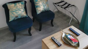 A room with two dark blue armchairs, a small wooden table with a remote control and magazines, and a wall-mounted light fixture above a decorative metal piece. - Studio Cosy & Cocon Bronze (Mulhouse)
