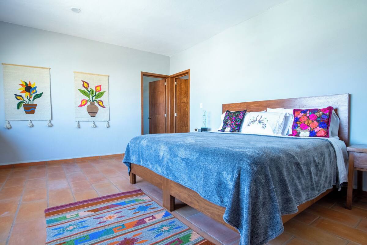 A bedroom with a bed, a wooden headboard, a colorful rug, and two wall-mounted paintings of potted plants.